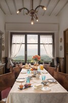 Comedor con una larga mesa dispuesta para una comida, ventanas grandes con cortinas y vistas al campo.