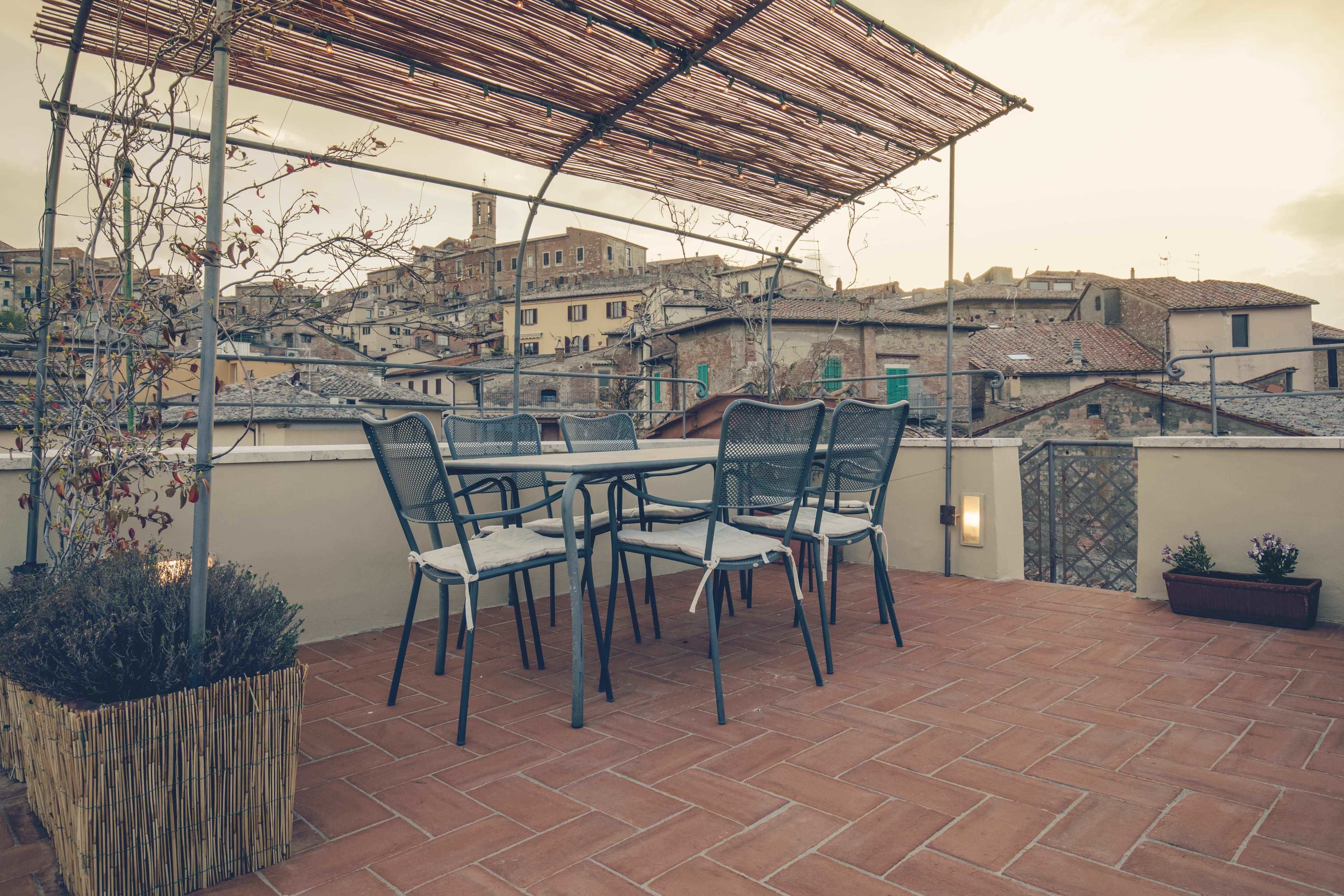 A terrace with a table and chairs, a woven basket, and a view of a town with buildings and a tower.