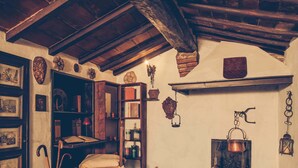 Una habitación rústica con suelo de piedra, vigas de madera, chimenea y estantes con diversos objetos.