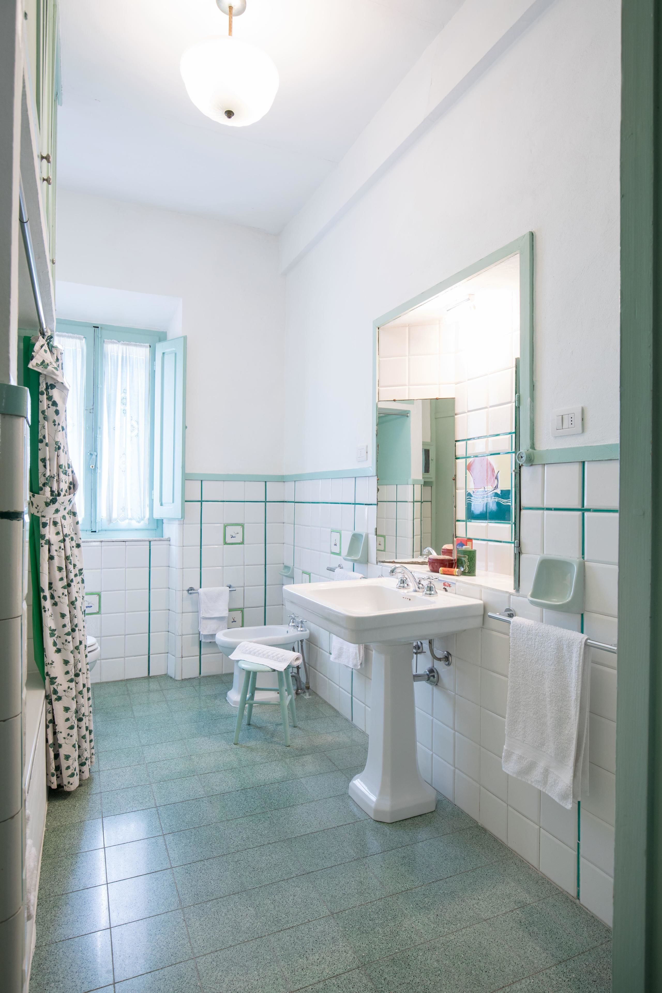 Ein Badezimmer mit einem weißen Waschbecken, Spiegel und Toilette. Es gibt ein Fenster mit Vorhängen und einen grün gefliesten Boden.