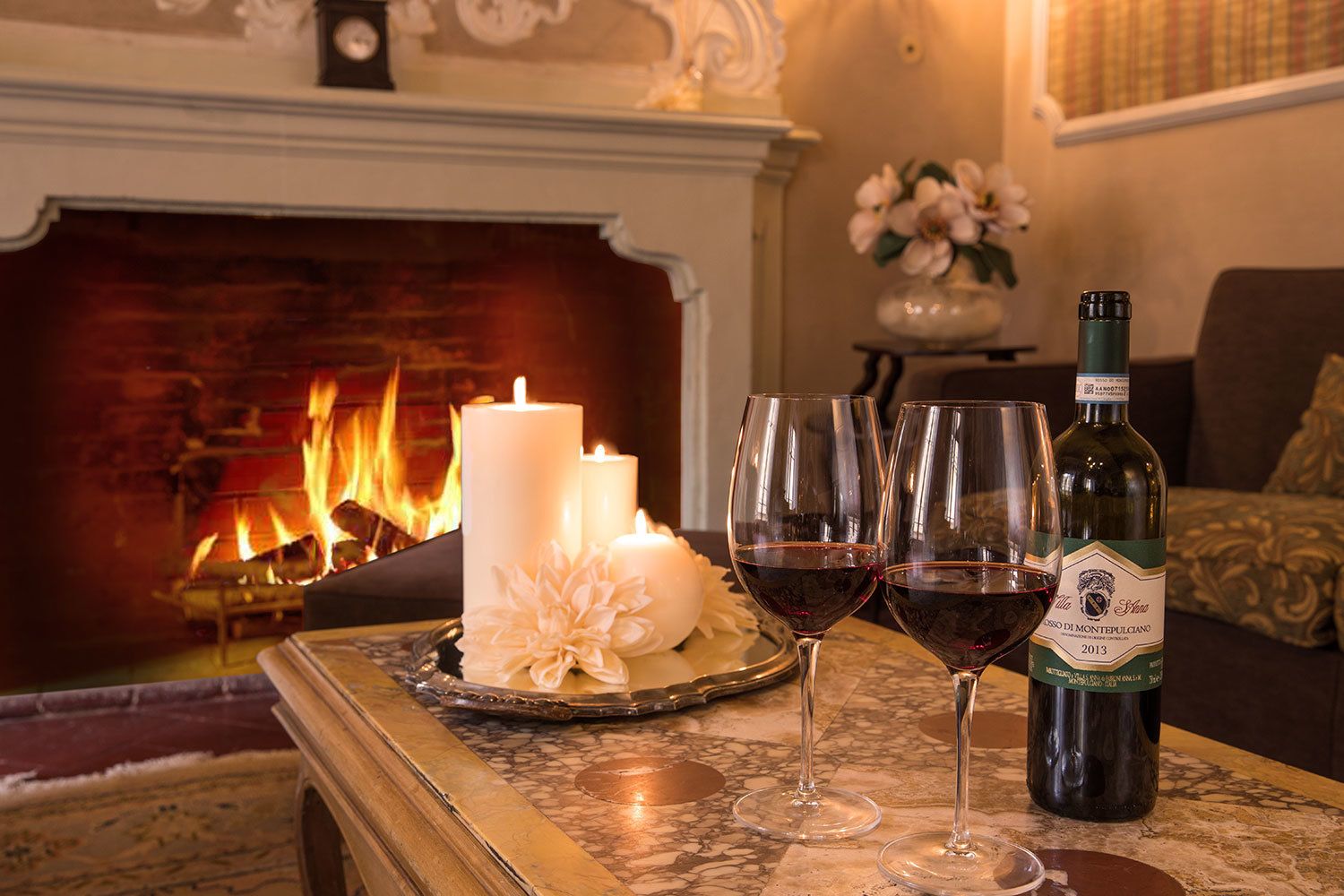 Une cheminée avec des bougies allumées, une bouteille de vin et deux verres à vin sur une table en marbre.