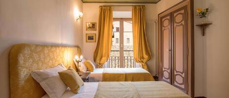 A hotel room with two beds, a balcony with yellow curtains, a wooden wardrobe, and a small shelf with a flower.