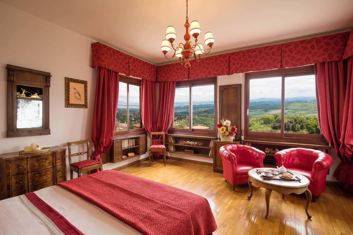 Una habitación con una cama grande, dos sillones rojos, una mesa de madera y vistas al campo a través de amplios ventanales.