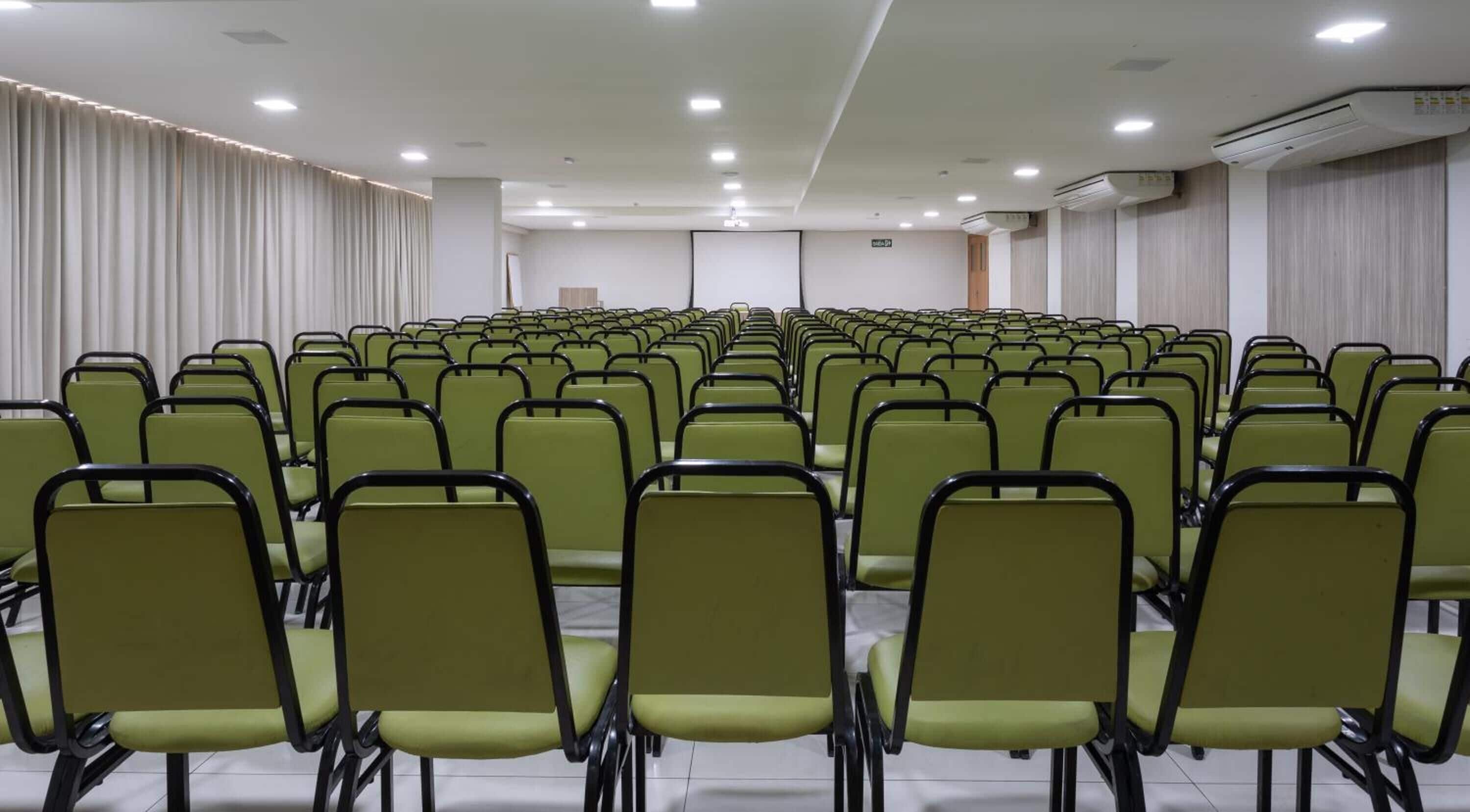 Sala de conferência com fileiras de cadeiras verdes e cortinas brancas.