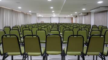 Sala de conferência com fileiras de cadeiras verdes e cortinas brancas.