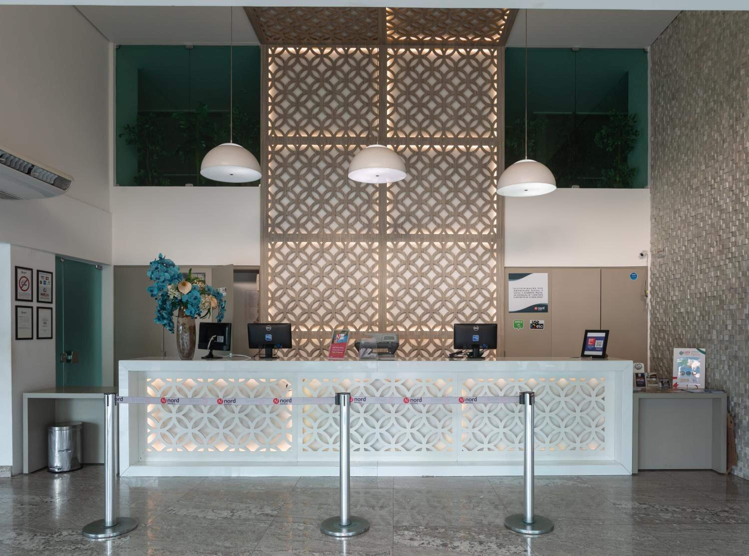 Uma área de recepção com divisória estampada, duas luminárias pendentes, um balcão de recepção com computadores e um arranjo floral azul decorativo.