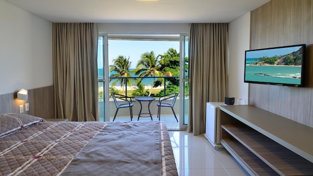 A hotel room with a large bed, a TV mounted on the wall, a balcony with a table and chairs, and a view of the ocean.