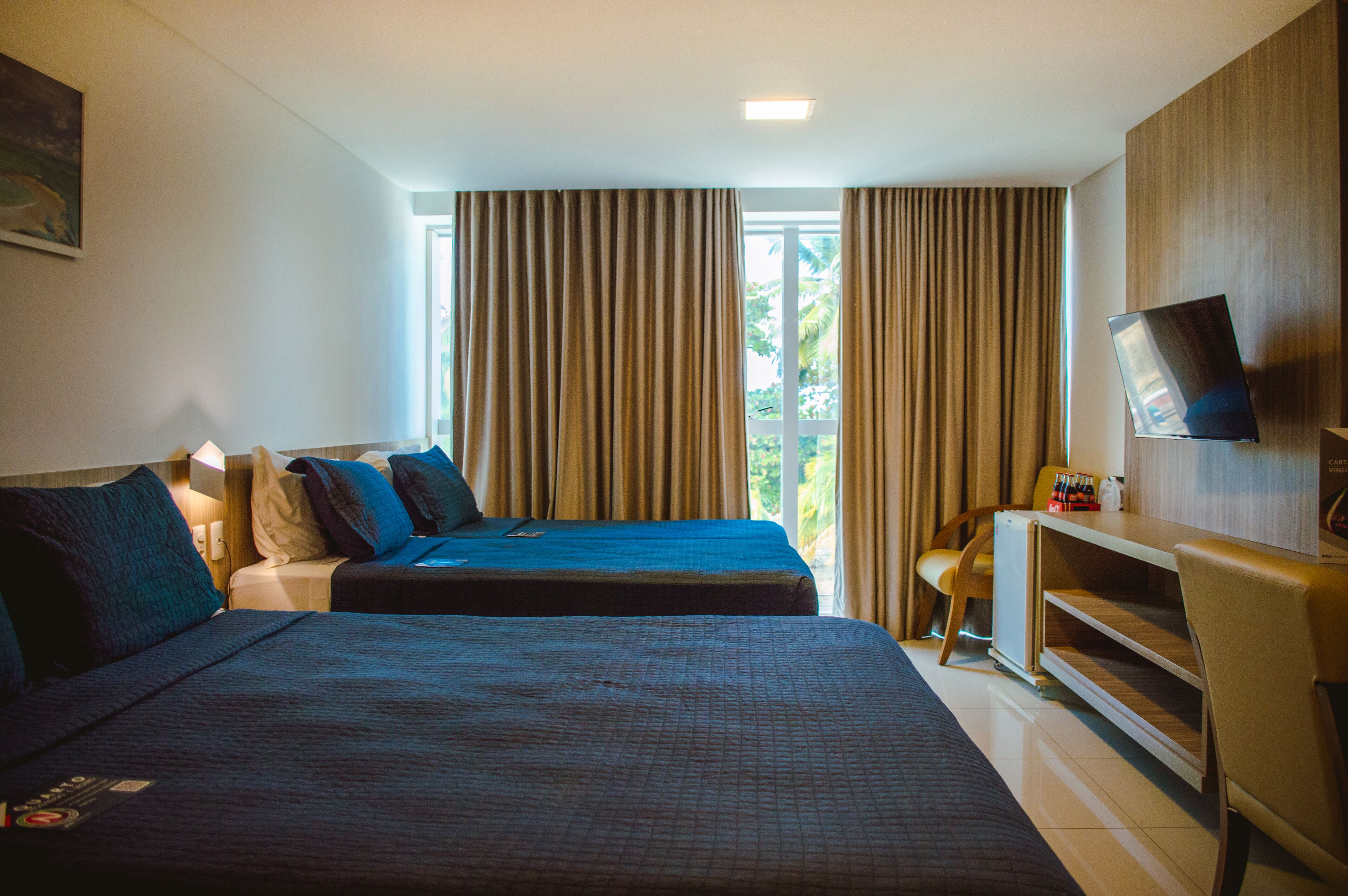 Quarto de hotel com duas camas, uma escrivaninha, televisão e janela com cortinas.