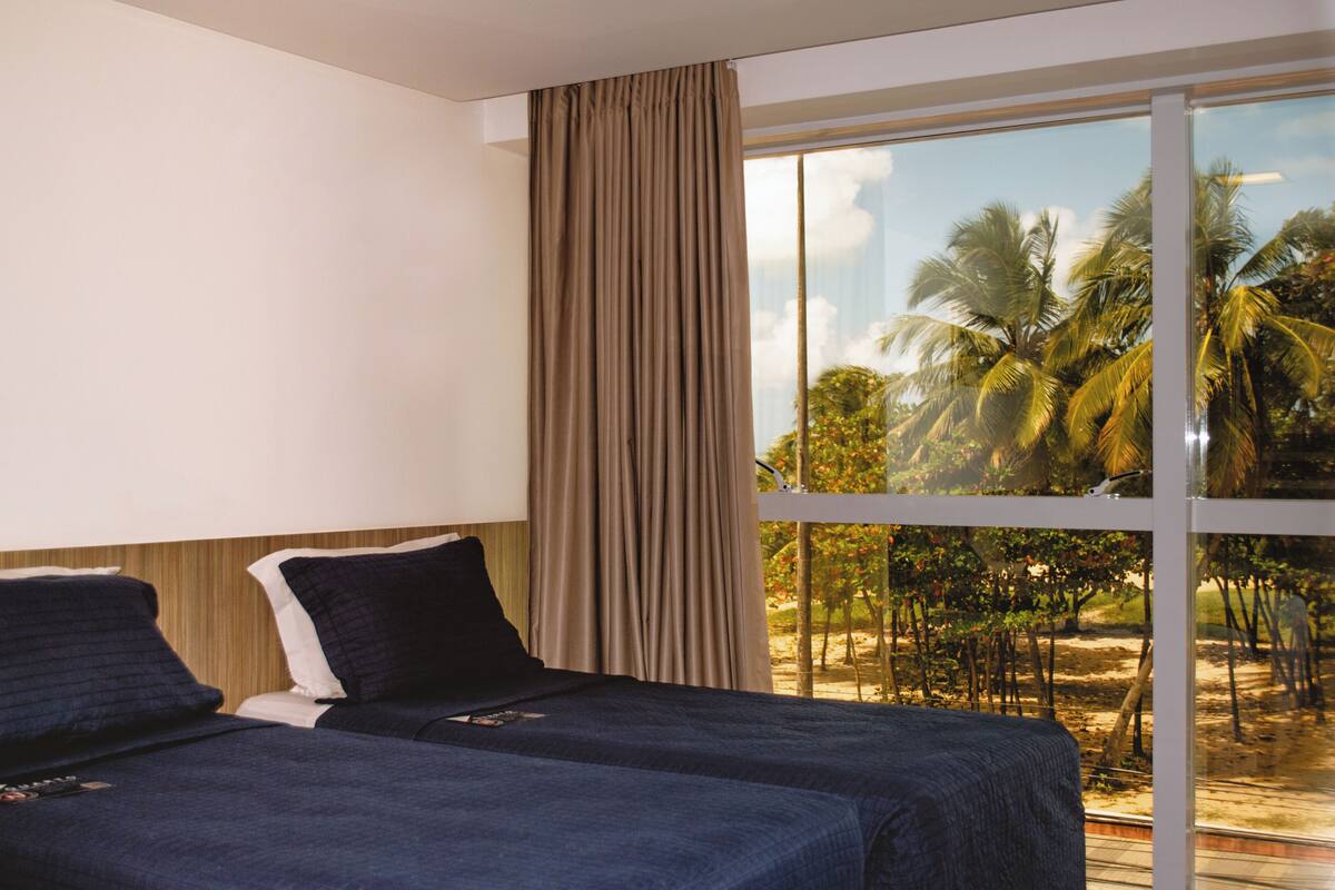 Una habitación de hotel con dos camas, un ventanal con vista a palmeras y una playa.