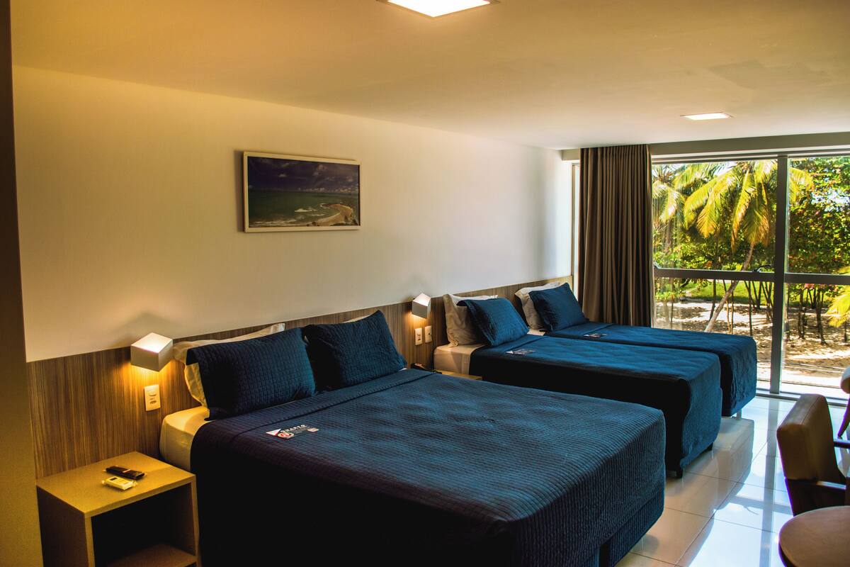 Habitación de hotel con dos camas, una mesita de noche, una lámpara, un cuadro en la pared y vistas a palmeras a través de la ventana.