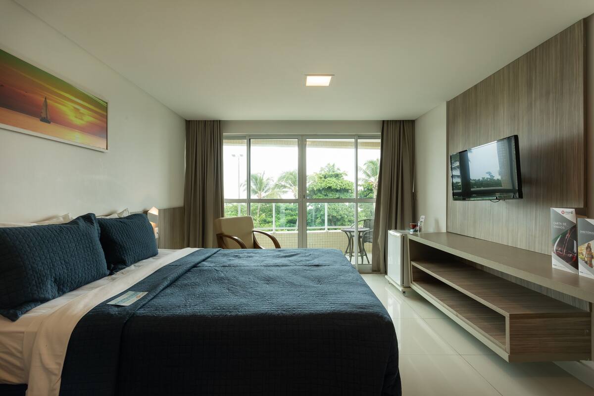 Habitación de hotel con una cama grande, un televisor montado en la pared, un escritorio con una silla y un balcón con vistas a palmeras.