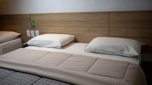Habitación de hotel con cabecera de madera, dos almohadas blancas, colcha de cuadros y una mesita de noche con enchufe verde.