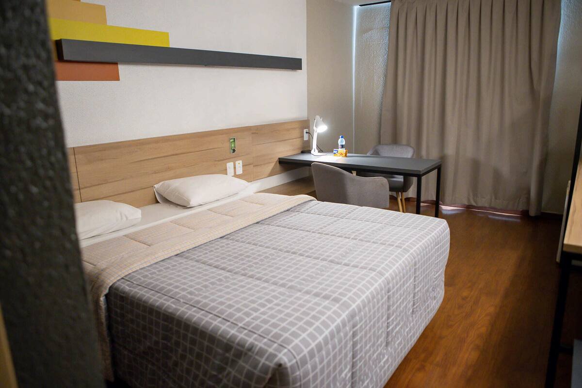 A hotel room with a bed, bedside table, chair, and a desk with a lamp.