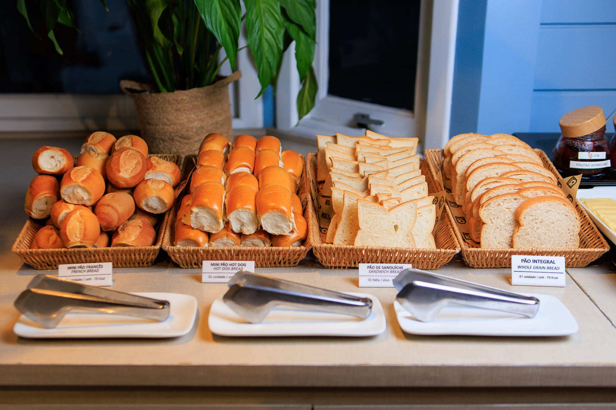A display of various bread types with labels, tongs, and a potted plant in the background.