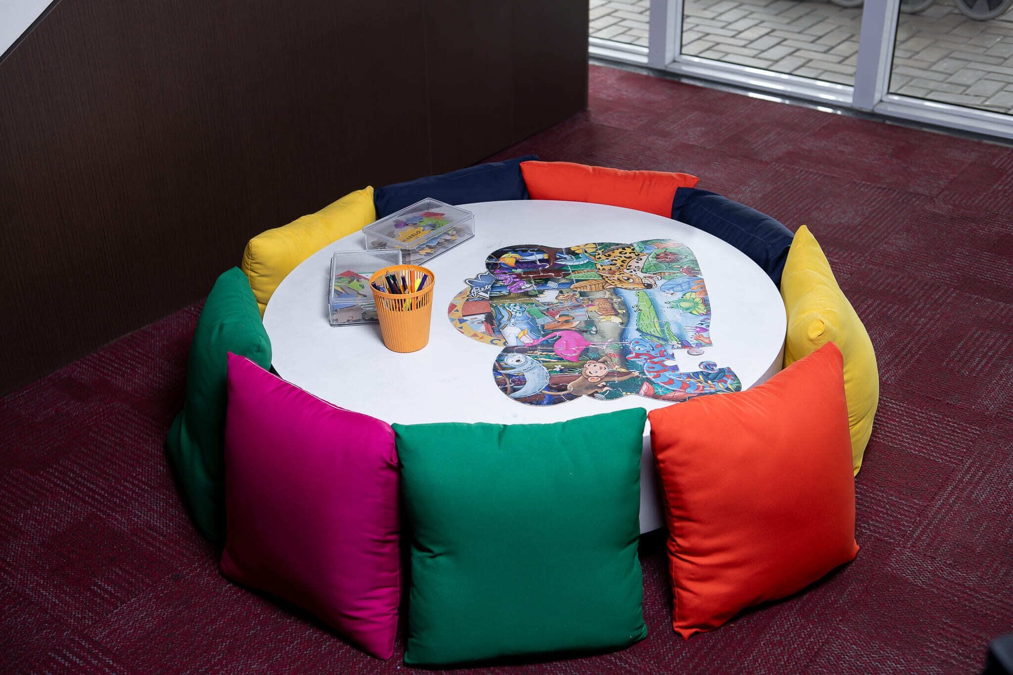A round table with colorful cushions and a puzzle in the center, surrounded by a red carpeted floor.