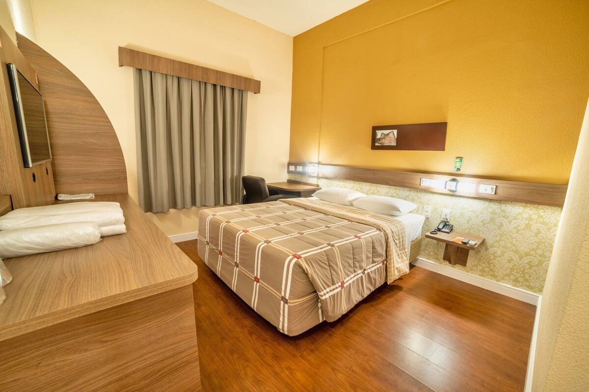 A hotel room with a bed, a desk, a chair, a nightstand, and a window with curtains.