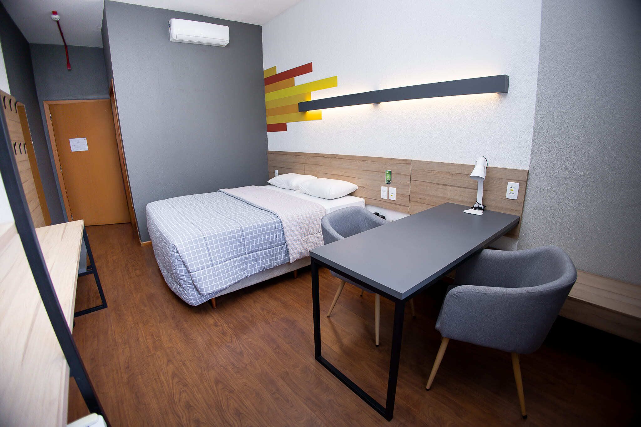 Quarto de hotel pequeno com uma cama, escrivaninha, cadeira, luminária na parede e porta.