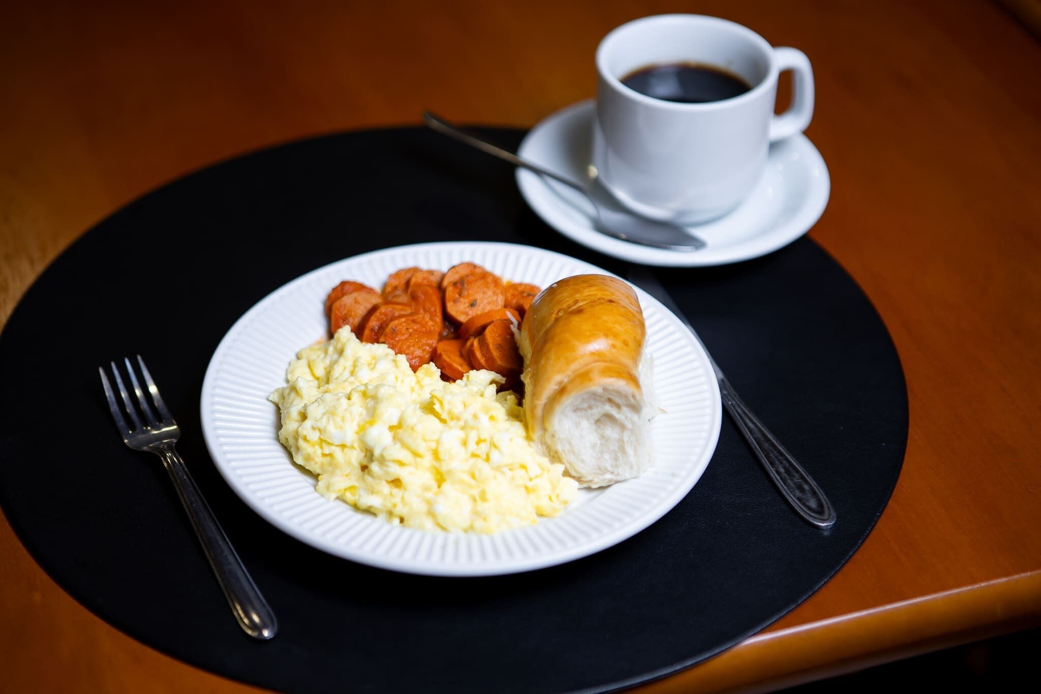 Um prato com ovos mexidos, salsicha e um croissant numa bandeja preta, acompanhado de uma chávena de café e uma colher.