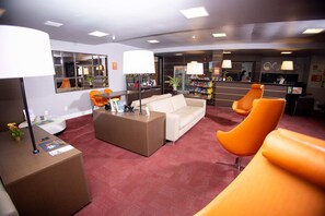 A hotel lobby with a reception desk, a sofa, orange chairs, a coffee table, and a large mirror.