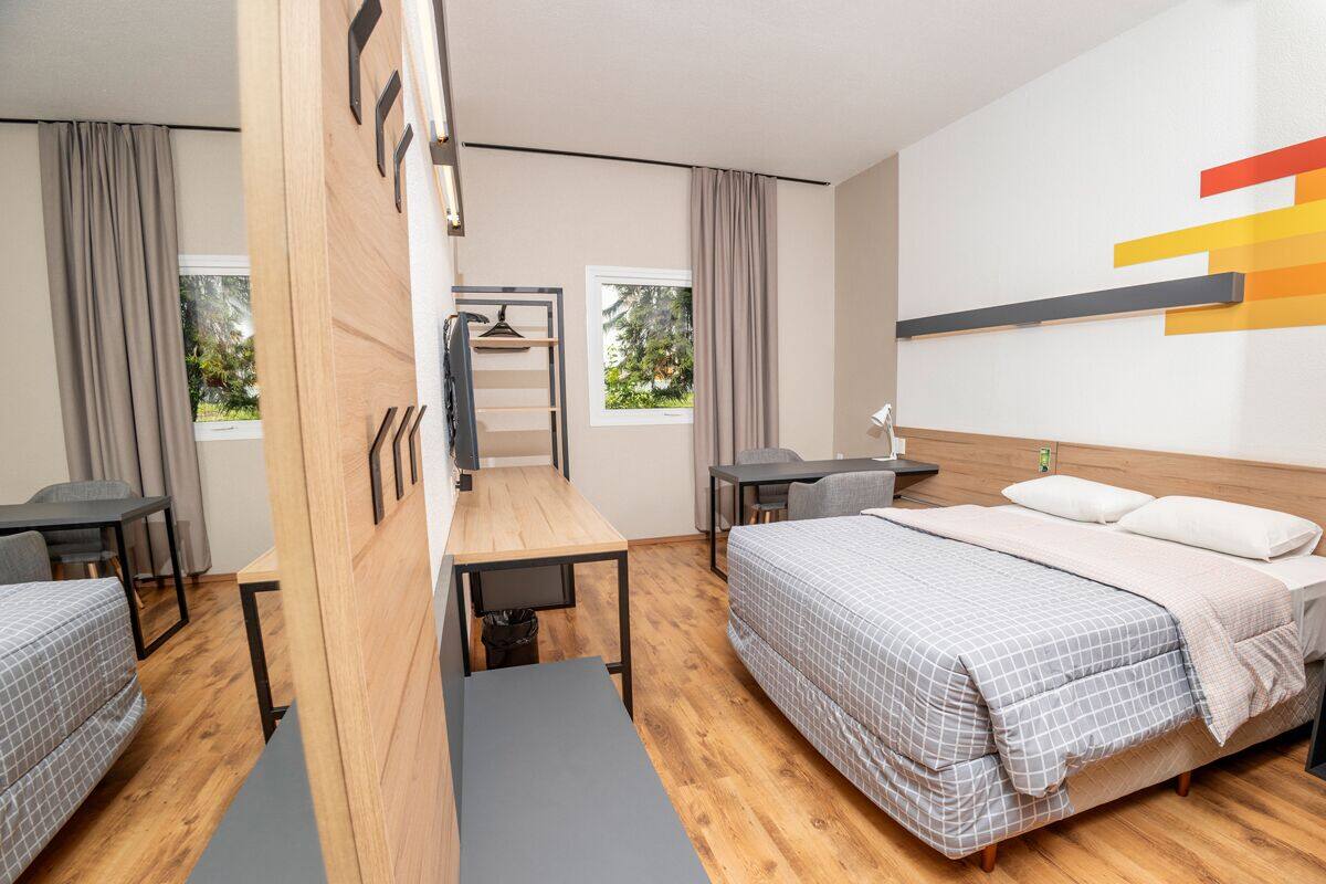 A modern hotel room with a wooden desk, two single beds, a large bed, a wardrobe, and a view of greenery through the windows.