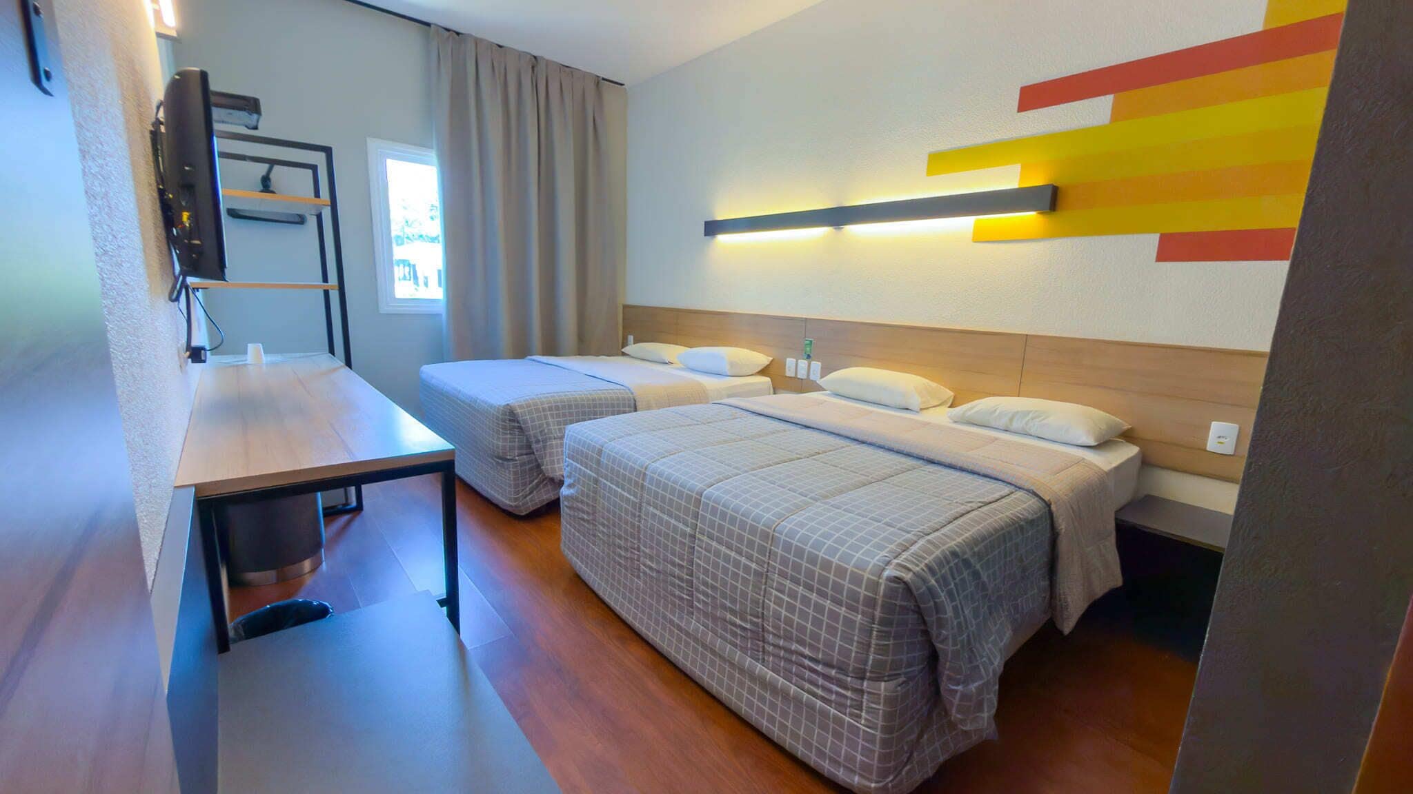 A hotel room with two beds, a desk, a chair, a TV, and a wall-mounted light fixture.