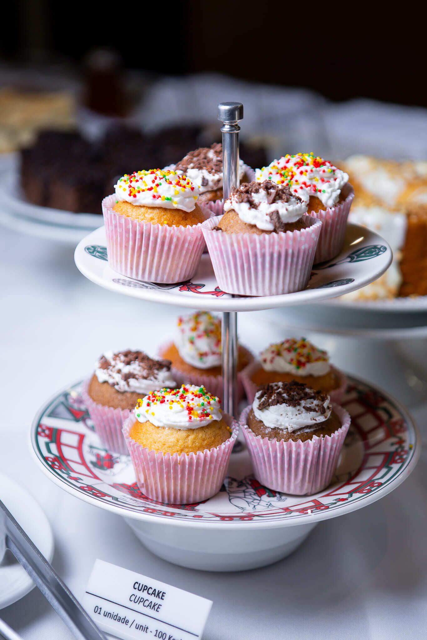 A tiered stand with cupcakes in pink paper cups, a metal stand, and a plate with a decorative border.