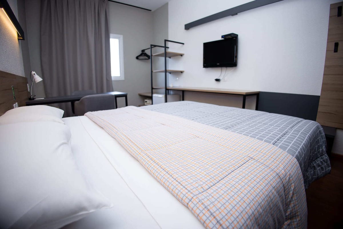 A hotel room with a bed, a desk, a chair, a TV, and a window with curtains.