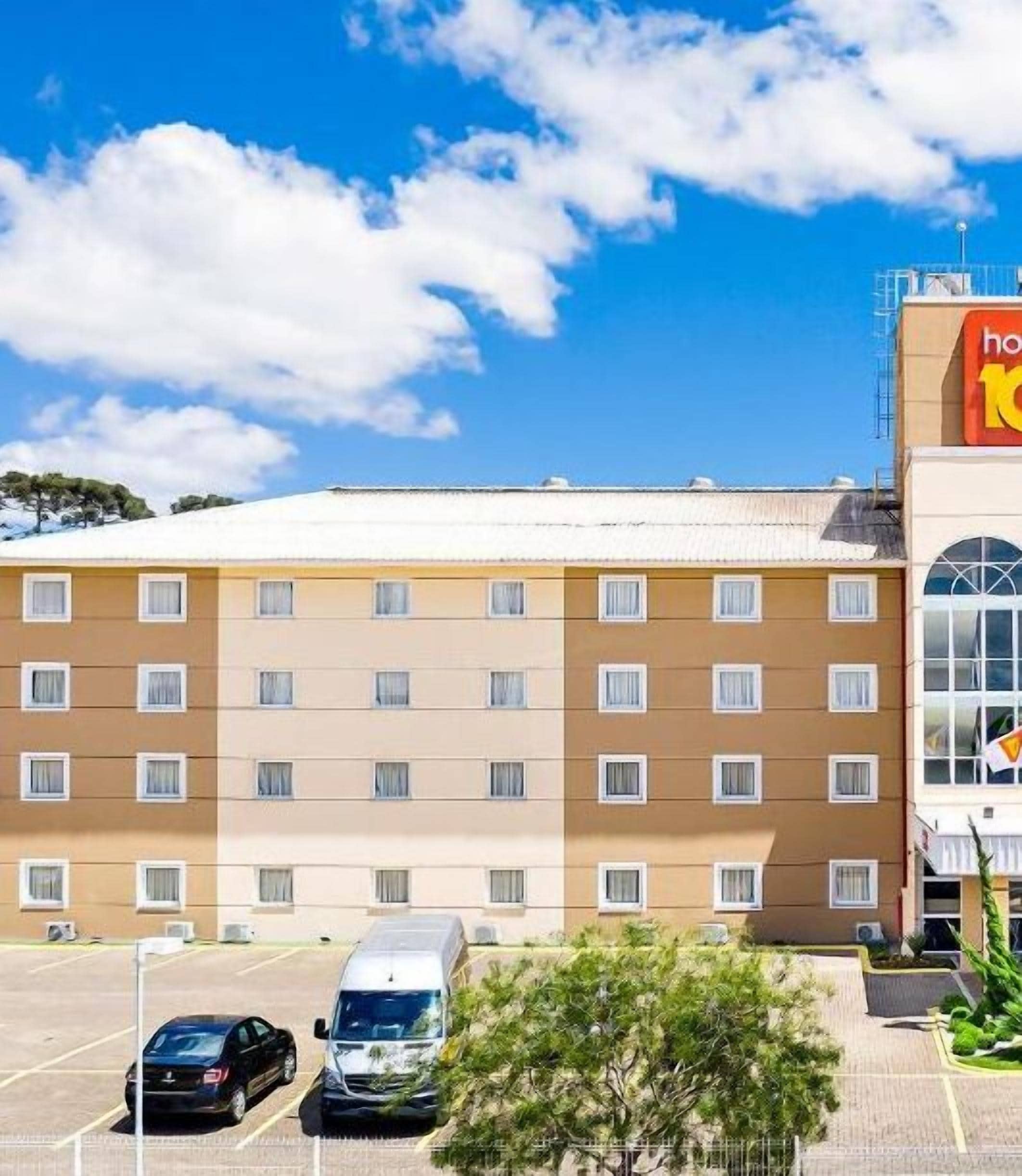 A multi-story hotel with a flat roof, a parking lot with several cars, and a clear blue sky with clouds.