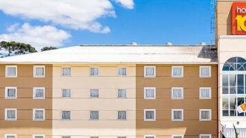 A multi-story hotel with a flat roof, a parking lot with several cars, and a clear blue sky with clouds.