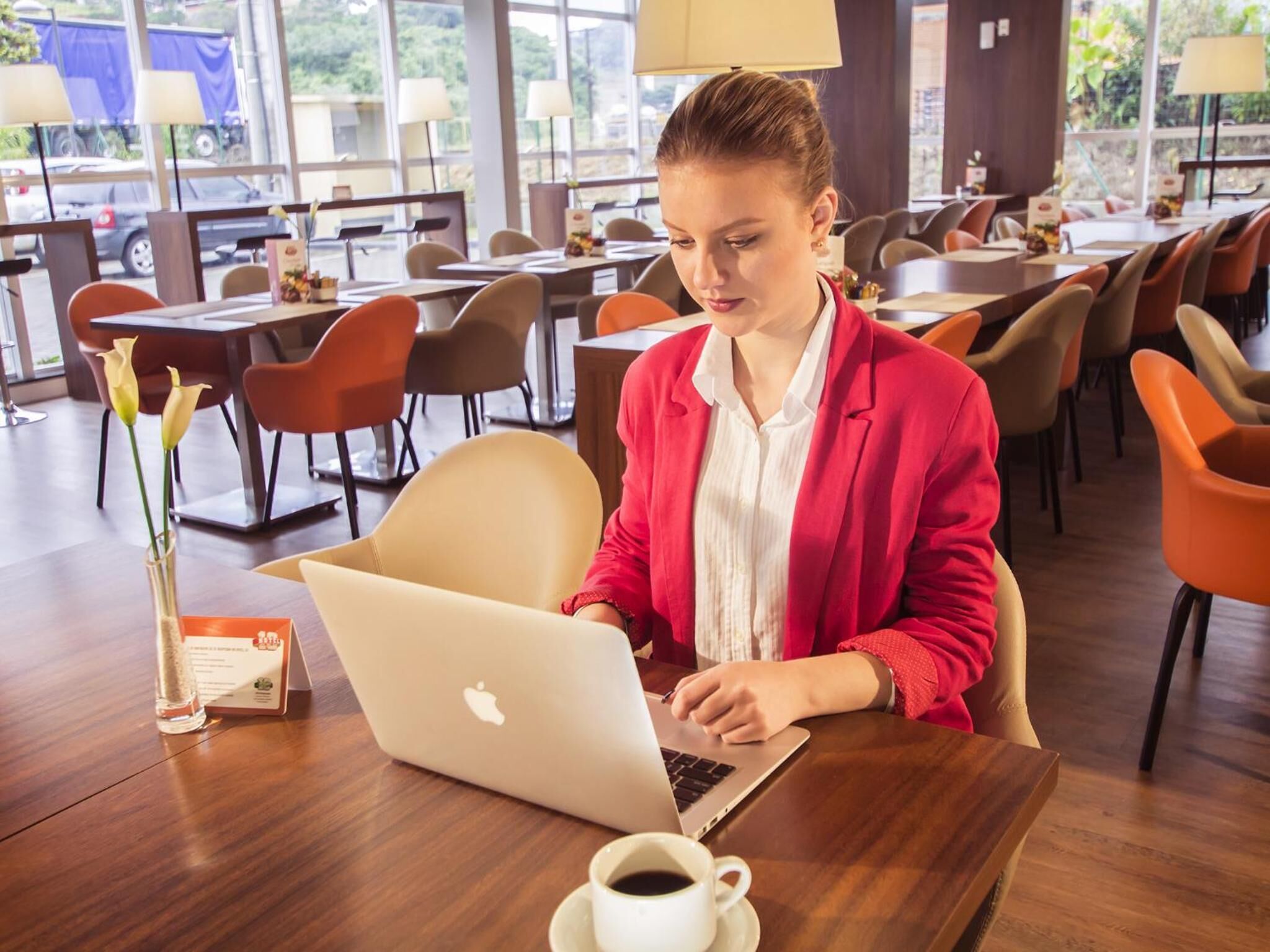 Uma pessoa usando um laptop em uma mesa em área de refeições moderna, com várias mesas e cadeiras.