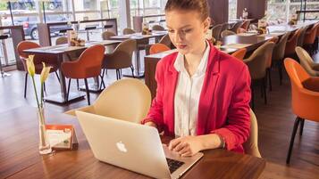 Uma pessoa usando um laptop em uma mesa em área de refeições moderna, com várias mesas e cadeiras.