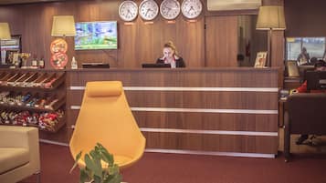 Zona de recepción de un hotel con un mostrador de madera, varios relojes que marcan diferentes husos horarios, una pantalla de televisión y un área de descanso con una silla amarilla y una pequeña mesa con una planta.