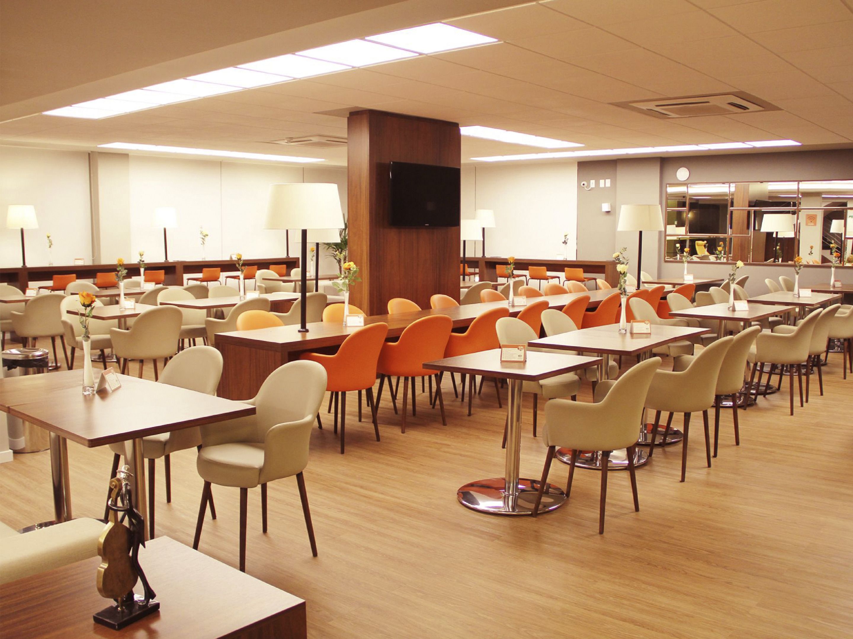 A spacious dining area with multiple tables and chairs, a central pillar, and a television mounted on the wall.