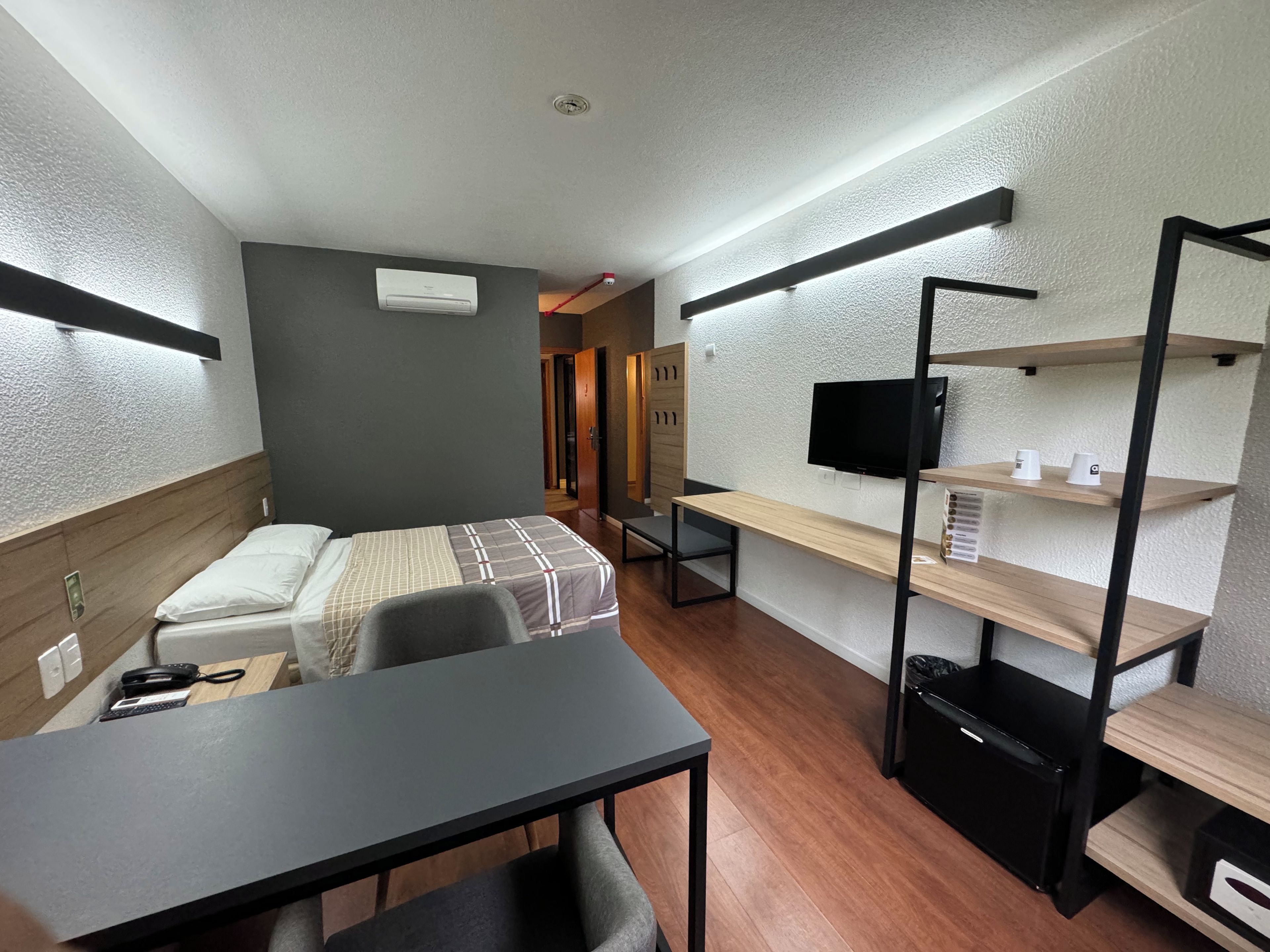 Quarto de hotel compacto com cama, escrivaninha, cadeira, TV e prateleiras embutidas.