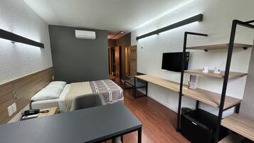 Quarto de hotel compacto com cama, escrivaninha, cadeira, TV e prateleiras embutidas.