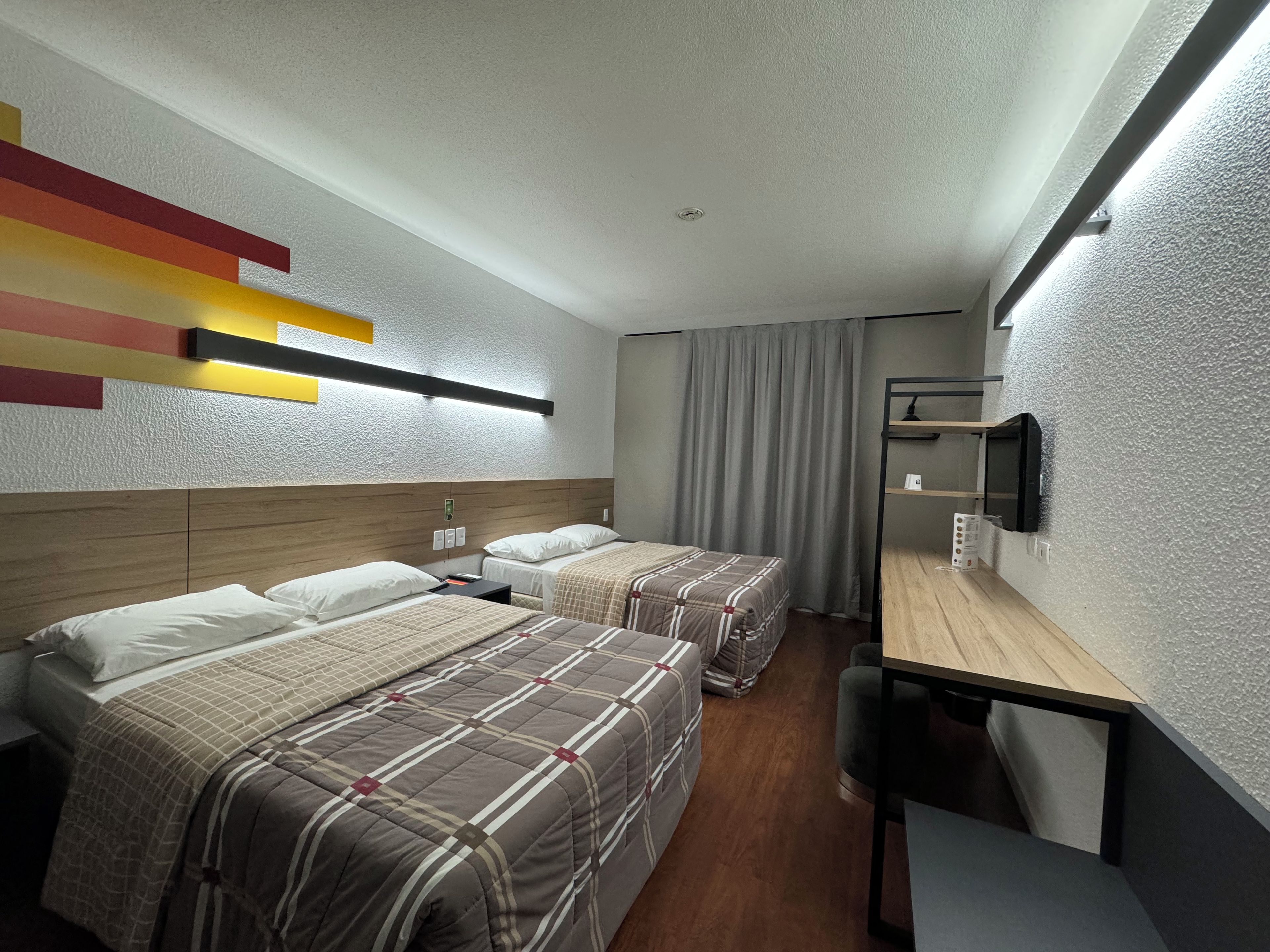 Quarto de hotel com duas camas, uma escrivaninha, televisão e luminária moderna.