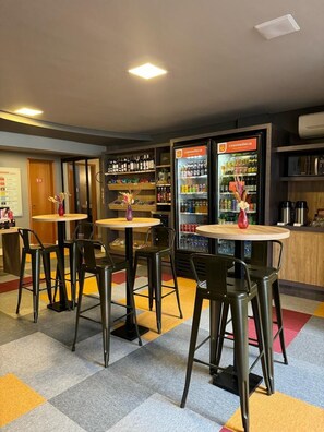 Un área de bar moderna con mesas redondas y sillas de metal negro, una estación de bebidas refrigerada y estantes llenos de botellas y aperitivos.