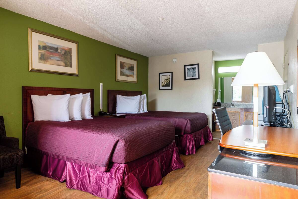 A hotel room with two beds, a desk, and a chair. There are framed pictures on the wall and a lamp on the desk.