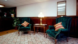 Eine Lobby mit zwei grünen Sesseln, einem kleinen Tisch mit Lampe, einem Topfpflanzen und einem gerahmten Verzeichnis an der Wand.