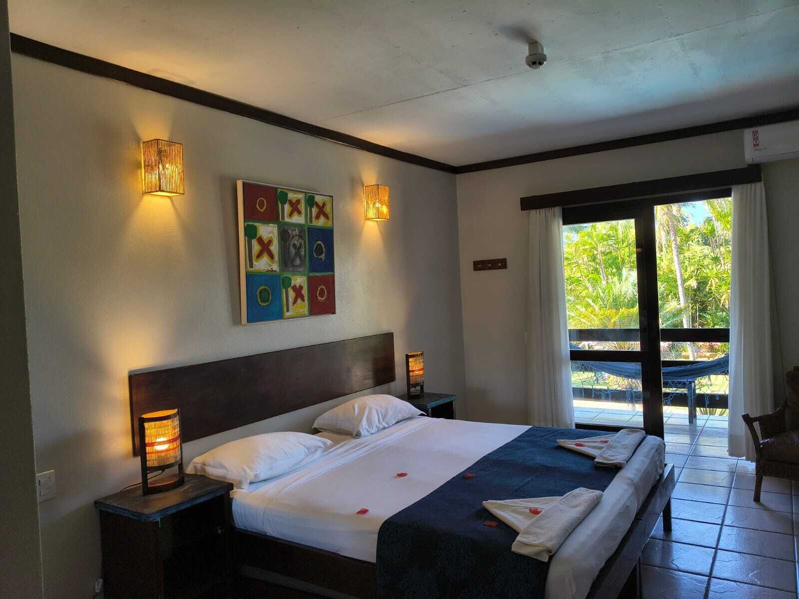 A hotel room with a large bed, two bedside lamps, a colorful artwork on the wall, and a view of the outdoors through a sliding door.