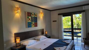 Quarto de hotel com uma cama grande, duas luminárias de cabeceira, uma obra de arte colorida na parede e vista para o exterior através de uma porta de correr.