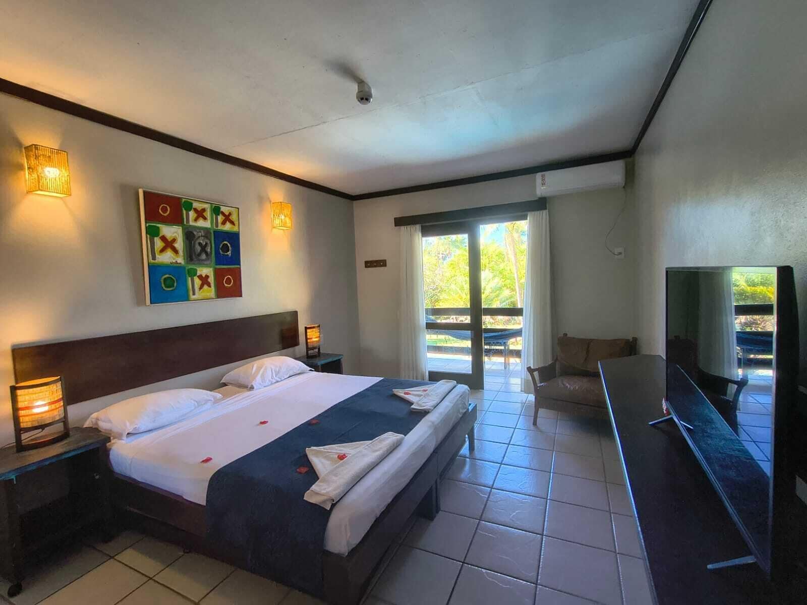A hotel room with a bed, bedside lamps, a television, a balcony with a hammock, and a colorful artwork on the wall.
