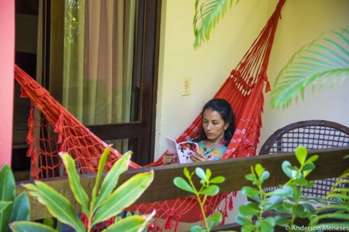 A person reading a book in a hammock on a balcony with plants and a chair.