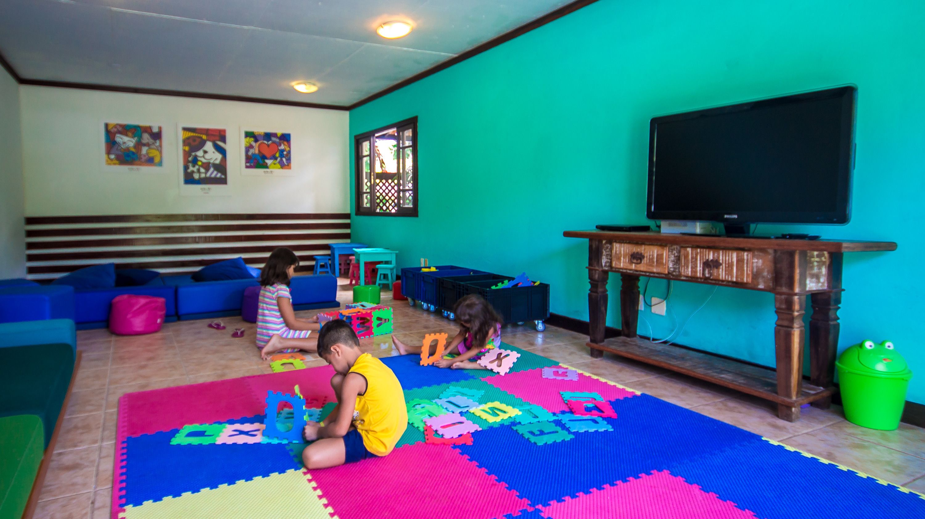 Quarto com tapetes de brincadeira coloridos, uma televisão e uma janela com treliça decorativa.