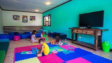 Quarto com tapetes de brincadeira coloridos, uma televisão e uma janela com treliça decorativa.