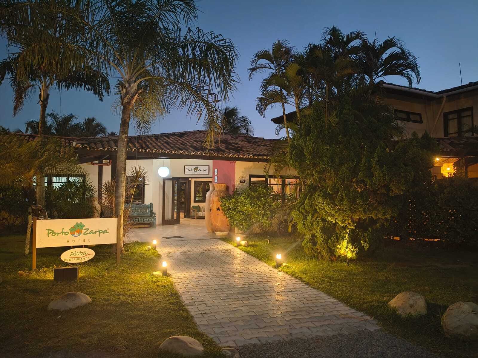 Entrada de hotel com um caminho pavimentado que leva à porta, cercada por palmeiras e uma placa iluminada.