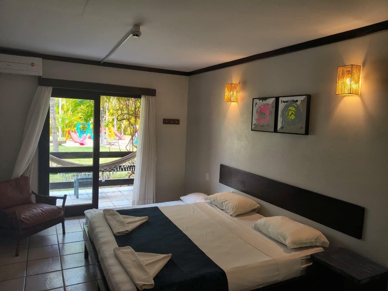 Quarto de hotel com cama, cadeira, mesa de cabeceira e vista para uma área de lazer ao ar livre.