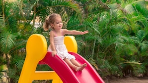 A child on a red slide in a playground with yellow and blue structures, surrounded by green foliage. - Porto Zarpa Hotel (Mata de São João)