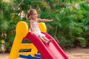 Un bambino su uno scivolo rosso in un parco giochi con strutture gialle e blu, immerso nel verde.