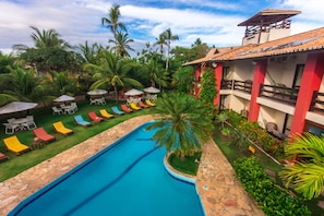 A pool area with lounge chairs and umbrellas, surrounded by palm trees and a building with a tiled roof. - Porto Zarpa Hotel (Mata de São João)